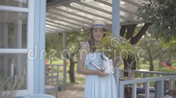 可爱的年轻女子戴着草帽穿着白色连衣裙微笑着在浇灌罐前嗅着野花视频的预览图