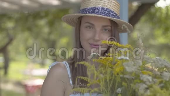 肖像可爱的年轻女子戴着草帽穿着白色连衣裙看着镜头微笑捧着野花视频的预览图