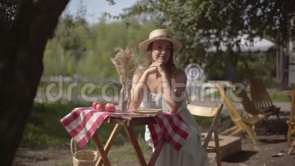 年轻的女孩戴着草帽和白色的连衣裙坐在小桌子旁看着相机农村生活方式休闲视频的预览图