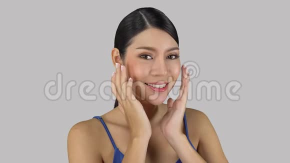 亚洲年轻女性在灰色背景下摆姿势快乐女性在脸上做轻按摩化妆概念视频的预览图