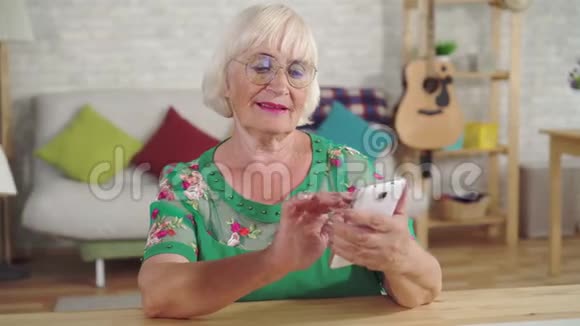 头发灰白的老年妇女使用智能手机坐在客厅的桌子旁视频的预览图