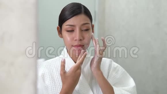 年轻可爱的微笑亚洲女人在她的浴室里做面部按摩在镜子前做美容视频的预览图