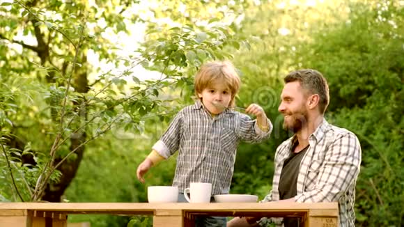 笑着可爱的小男孩坐在高椅上在绿色的自然背景下吃东西美国的花园派对父亲视频的预览图