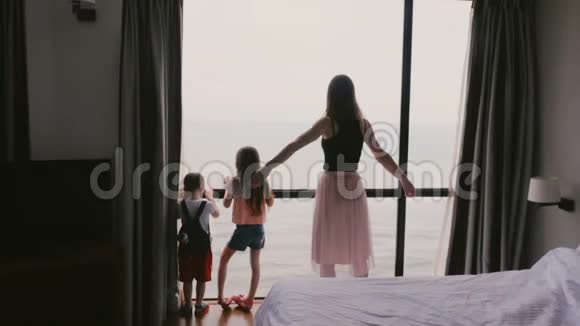 年轻快乐的母亲和两个小孩子一起跳在大窗户上玩得开心史诗般的海景慢动作视频的预览图