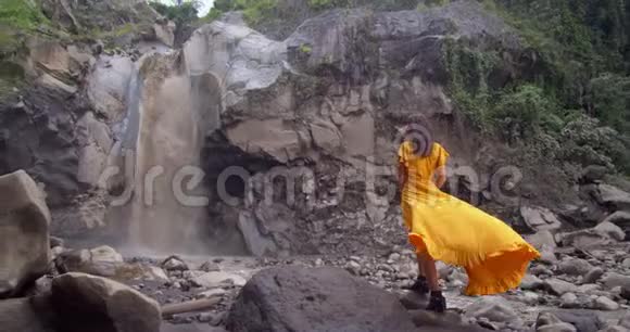 穿着黄色长裙的女人欣赏高山瀑布急速的海水从岩石中坠落大自然的可怕力量视频的预览图