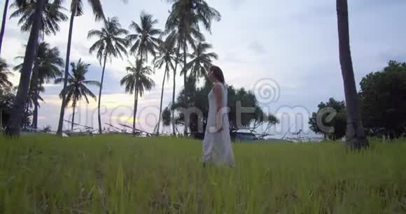 在温暖的夜晚穿着白色长裙的年轻女子在热带树木中漫步享受着她的时光视频的预览图