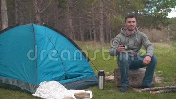 一个年轻人坐在森林里一个蓝色帐篷附近的大木头上用保温瓶喝茶视频的预览图