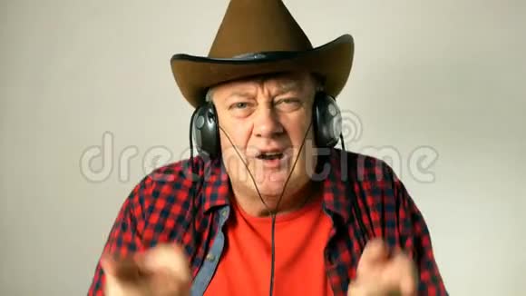 戴着牛仔帽的有表现力的成年老人听着耳机上的音乐边唱边跳边拍音乐视频的预览图