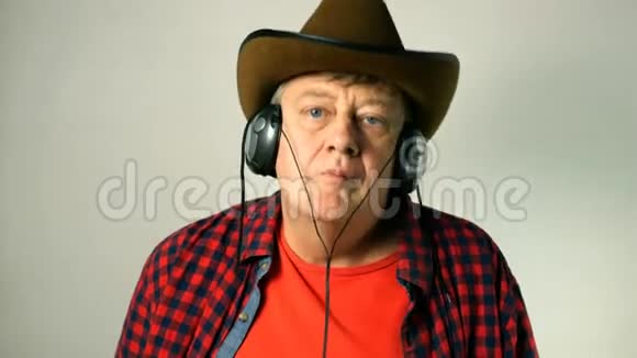 戴着牛仔帽的有表现力的成年老人听着耳机上的音乐边唱边跳边拍音乐视频的预览图