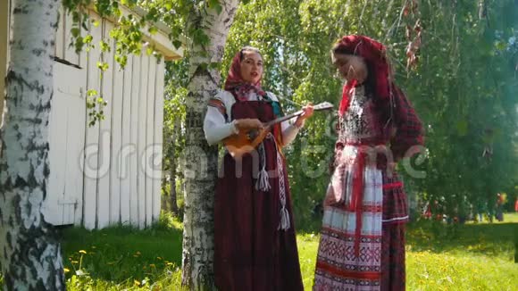 两个穿着俄罗斯服装的年轻女子站在草地上其中一个扮演巴拉莱卡视频的预览图