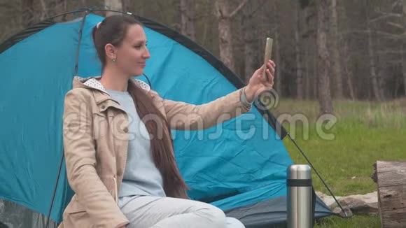 一个留着长发的年轻女孩正坐在树林里的一个蓝色帐篷附近通过电话中的视频链接交谈视频的预览图