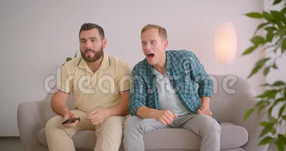 两名白人男子在电视上观看体育节目并在家里一起坐在沙发上庆祝视频的预览图