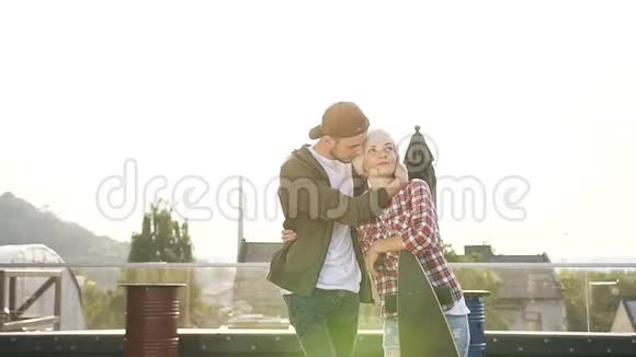 年轻迷人的一对相爱的夫妇在日落时在露台现代建筑的屋顶上带着滑板散步那个家伙视频的预览图