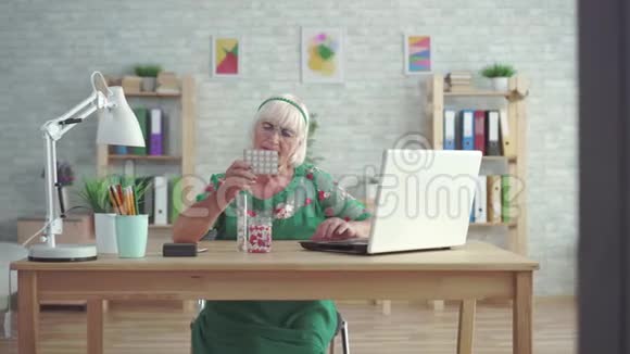 老妇人正在看一台笔记本电脑上有关坐在桌子旁的药片的信息视频的预览图