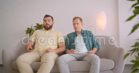 两名白人男子在电视上观看足球和在家中坐在沙发上庆祝的特写照片视频的预览图