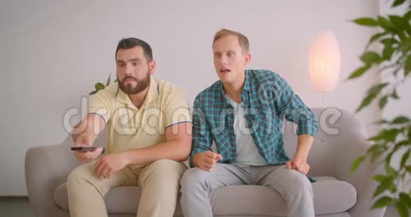 两位白种人男性朋友在电视上看体育节目一起坐在家里的沙发上庆祝视频的预览图