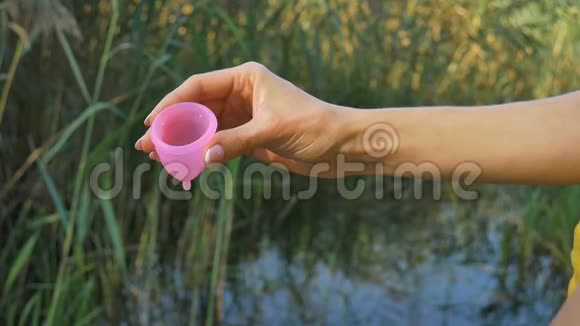 女人拿着一个粉红色的月经杯站在绿色的自然背景下与湖女性卫生概念视频的预览图