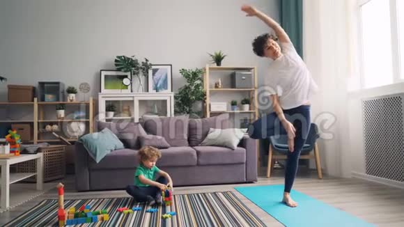 小男孩在地板上玩玩具时他运动的妈妈在垫子上做瑜伽视频的预览图