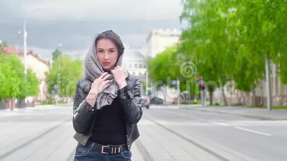 城市背景下穿黑色皮夹克的性感女孩的肖像手拿着灰色的围巾戴在头上夏日阳光明媚视频的预览图
