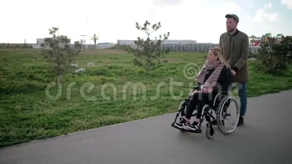 男人和年轻的金发女人坐在轮椅上一起在公园里散步视频的预览图