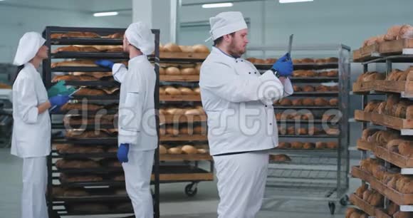 在一家面包店里主要的面包师带着一块新的平板电脑拍了一些新鲜烘焙面包的照片另外两个面包师在上面视频的预览图