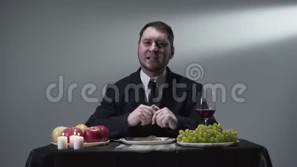 严肃的绅士穿着西装坐在小桌前安静地吃着肉葡萄苹果红酒和蜡烛视频的预览图