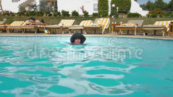 戴着黑帽子的白人老年女性在酒店的蓝色游泳池游泳在沙滩椅的背景下视频的预览图