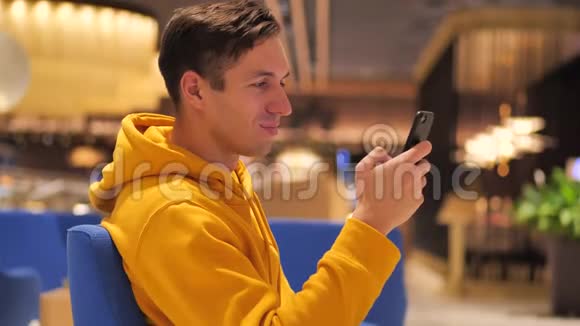 穿着黄色连帽衫的年轻人坐在咖啡馆购物中心使用现代智能手机视频的预览图