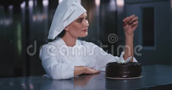 年轻可爱的面包师女士戴着厨师帽轻轻地把樱桃放在蛋糕上对着镜头微笑视频的预览图