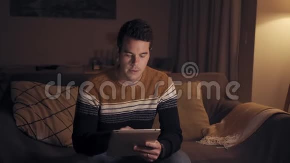 客厅深夜使用数码平板电脑坐在沙发上舒适的男人的肖像视频的预览图