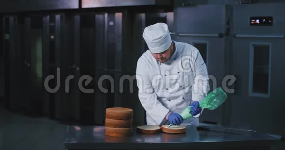 一个留着胡须的大个子男人戴着面包师的制服蓝色手套和厨师的帽子用白色绒毛装饰蛋糕的内部视频的预览图