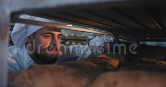面包厂的面包师有胡子的人在工业货架上拿一个新鲜的烤面包去食品工业视频的预览图