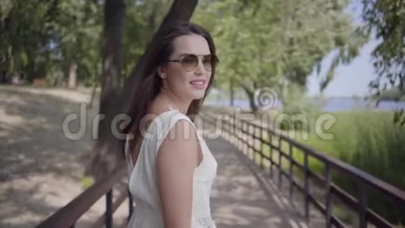 一位美丽的黑发少女戴着太阳镜穿着白色的夏季时装长裙走在木桥上视频的预览图