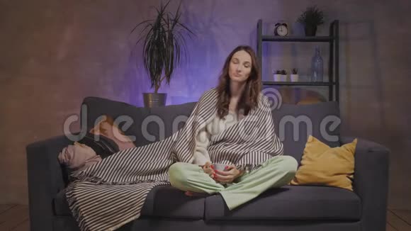 一个年轻的女人坐在一个舒适的房间里裹着毯子喝茶看电视视频的预览图
