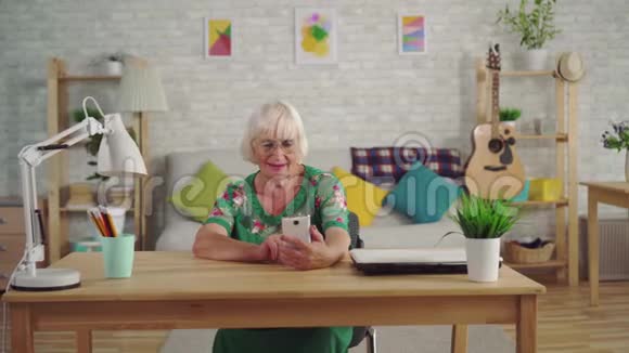 头发灰白的女性养老金领取者使用智能手机坐在客厅的桌子旁视频的预览图