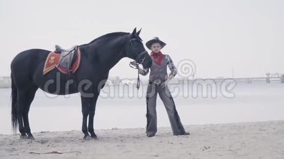 戴着帽子的金发女牛仔和她的马站在河滩上4K视频的预览图