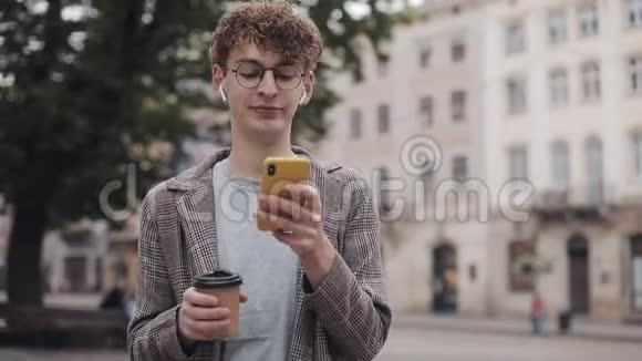 戴眼镜和无线耳机的年轻嬉皮士学生用手机微笑喝酒穿夹克视频的预览图