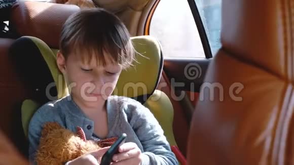 快乐可爱的小男孩在汽车儿童安全座椅上使用智能手机娱乐应用程序在阳光明媚的日子看着窗外视频的预览图
