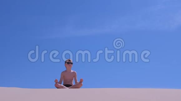 戴墨镜的小男孩正在天空背景下的海滩上沉思莲花姿势视频的预览图