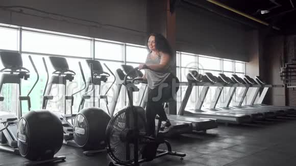 一个在健身房骑自行车的快乐大码女人的完整镜头视频的预览图