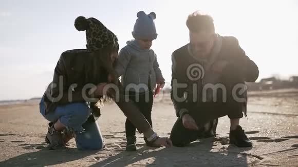 年轻的父母带着孩子蹒跚学步的男孩在寒冷多云多风的天气里玩沙子男人和女人帮助小男孩视频的预览图