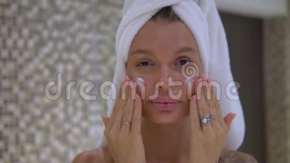 一位身材矮小可爱的年轻女士头上戴着白色毛巾早上在浴室里用化妆品按摩脸部视频的预览图