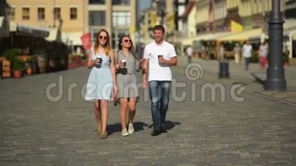 三个快乐的朋友一边喝咖啡一边在城市里走来走去两个女孩和一个男孩的全高画像视频的预览图