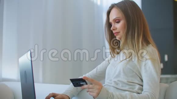 漂亮女孩正在网上付款拿着银行卡用现代笔记本电脑在家里坐在沙发上微笑财务视频的预览图