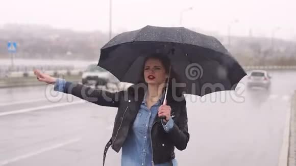 在大雨中一个漂亮的女孩在路上的伞下试图抓住一辆车视频的预览图