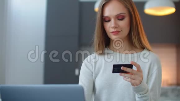 漂亮女孩正在网上付款拿着银行卡用现代笔记本电脑在家里坐在沙发上微笑财务视频的预览图