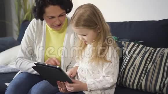 母亲或祖母坐在沙发上向小女孩展示数字技术视频的预览图