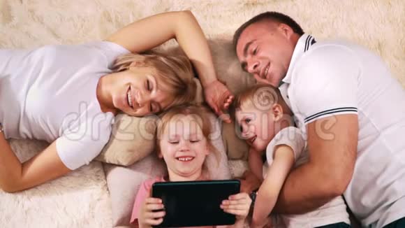 一个年轻的家庭在休息日躺在床上微笑着和孩子们一起看着平板电脑视频的预览图