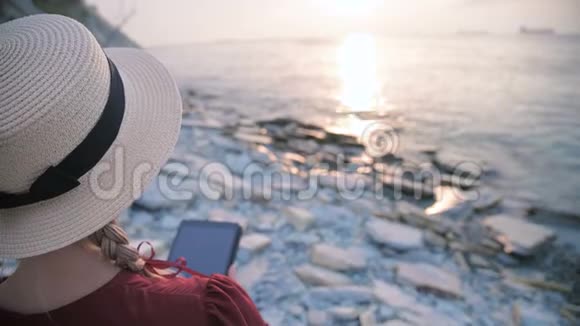 从后面可以看到一个漂亮的年轻女孩她穿着一件夏天的红色连衣裙和一顶草帽坐在海边的石头上视频的预览图