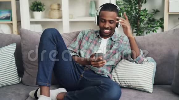 穿休闲服装的美籍非裔美国人听着耳机里的音乐坐在沙发上唱歌视频的预览图